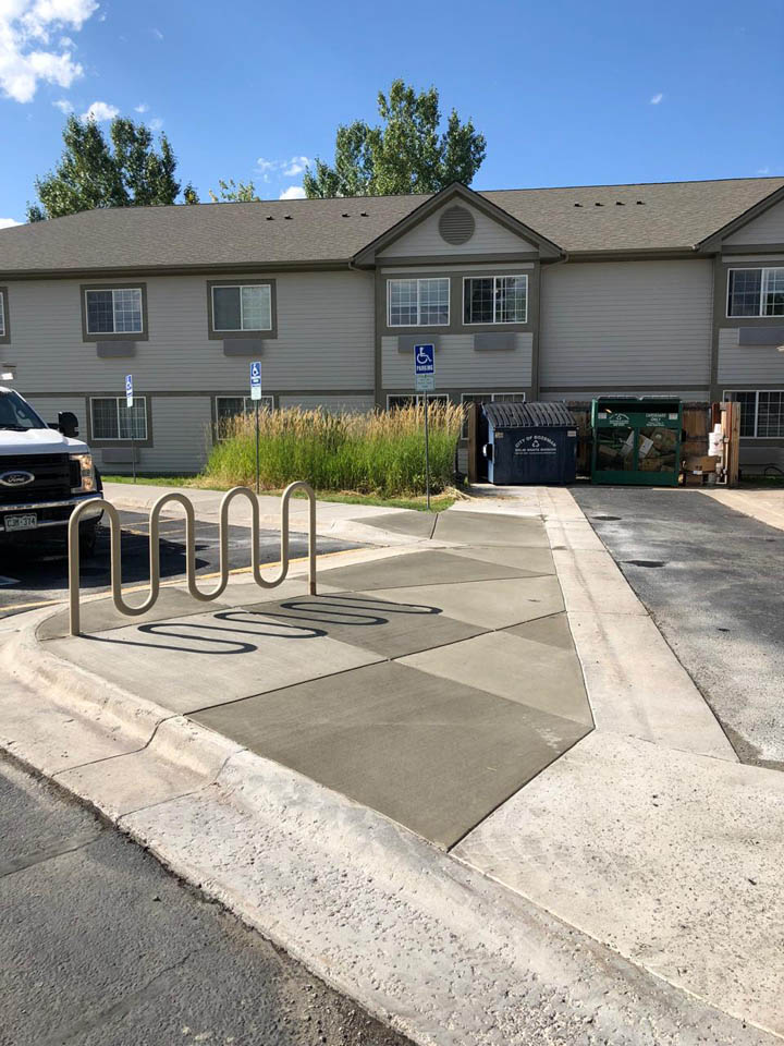 A sidewalk with a bike rack ends abruptly in front of a grassy area, blocking wheelchair access to two accessible parking spots near an apartment building. A dumpster is nearby.