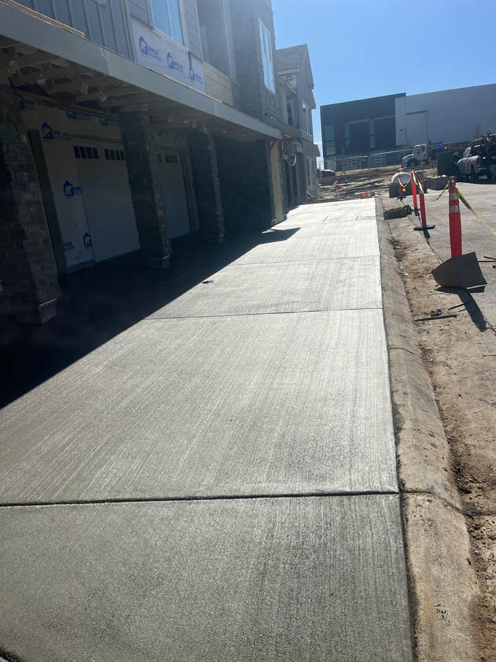 Freshly poured concrete driveway outside a house under construction, with traffic cones, construction equipment, and a modern building visible in the background under a clear blue sky.