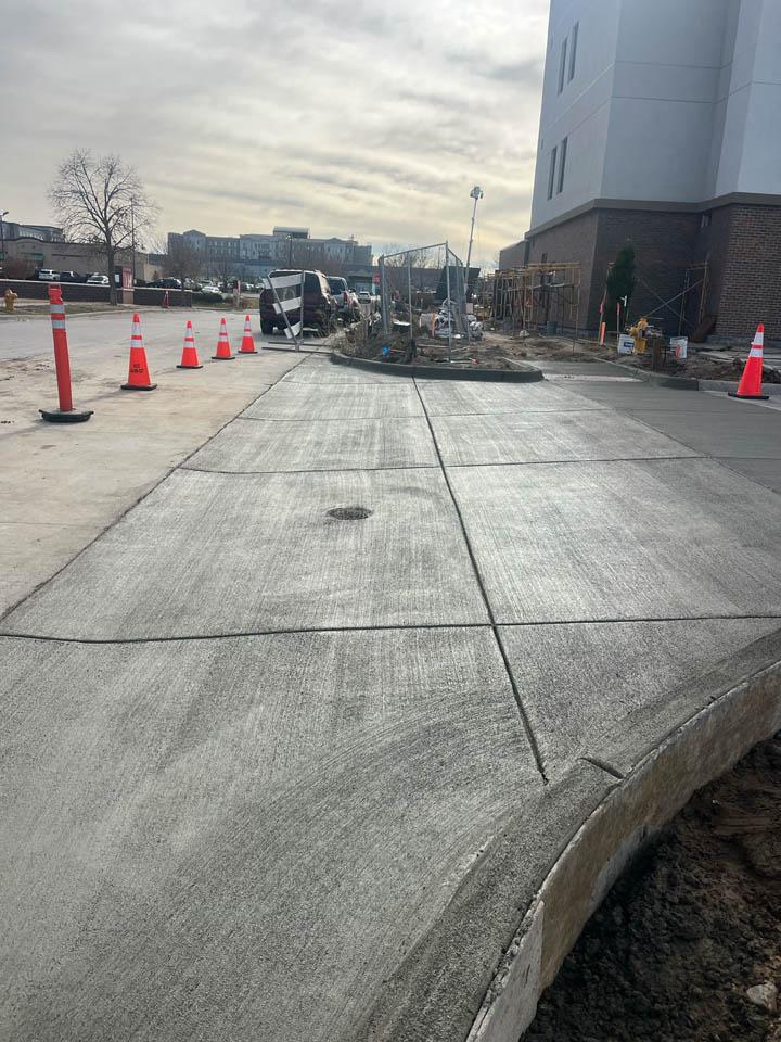 Freshly poured concrete sidewalk curves beside a building under construction. Orange traffic cones line the edge, and construction materials and workers are visible in the background under a cloudy sky.