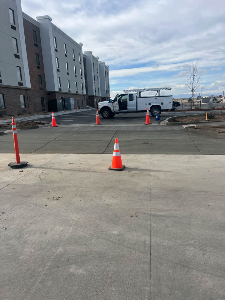 A white utility truck is parked on a freshly paved area near a modern multi-story building. Orange traffic cones block off the new pavement under a partly cloudy sky.