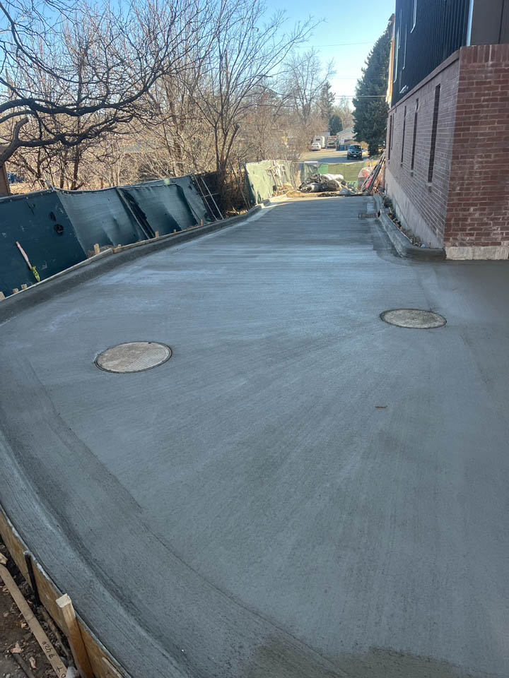 Freshly poured concrete driveway with two round utility covers, bordered by bricks on one side and a fence and trees on the other, with a house and street visible in the background.