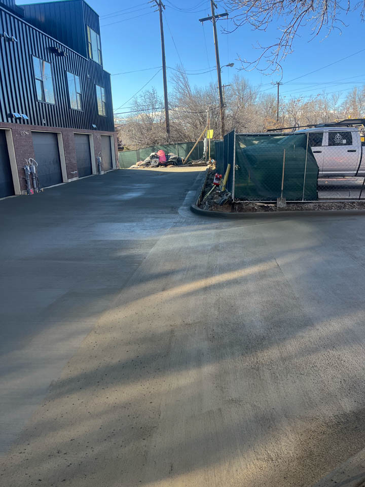 Freshly poured concrete pavement in front of a building with garages, surrounded by construction fencing; workers and construction materials are visible in the background under a clear blue sky.