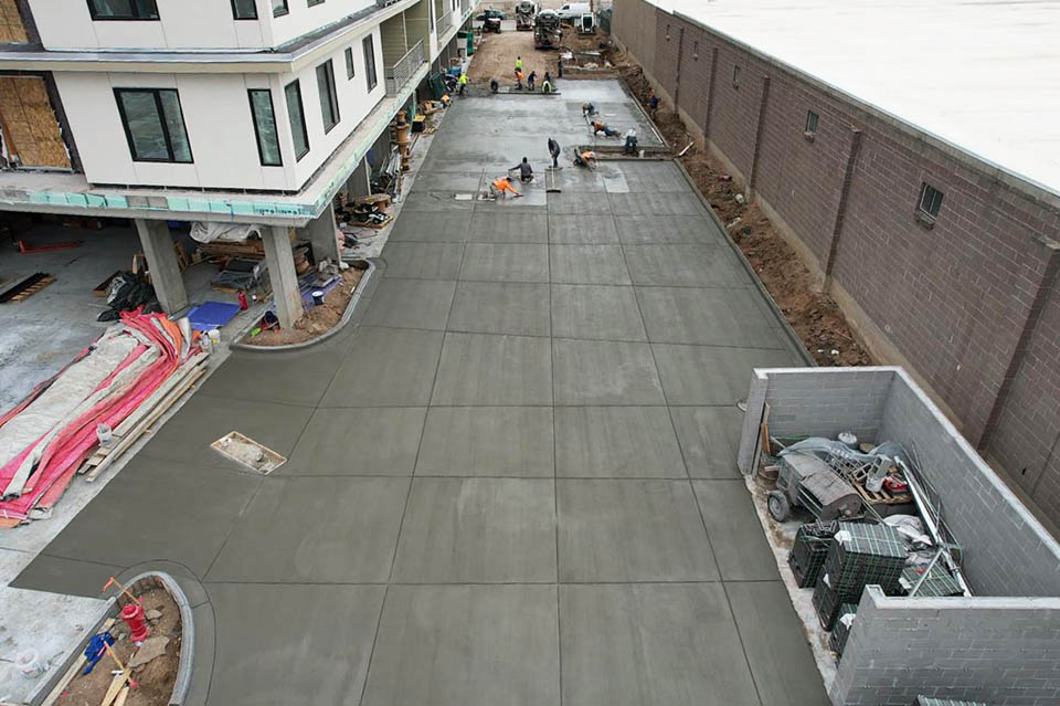 A group of construction workers smooth and finish freshly poured concrete in a large driveway area beside a modern building and a tall brick wall. Construction materials and equipment are scattered nearby.