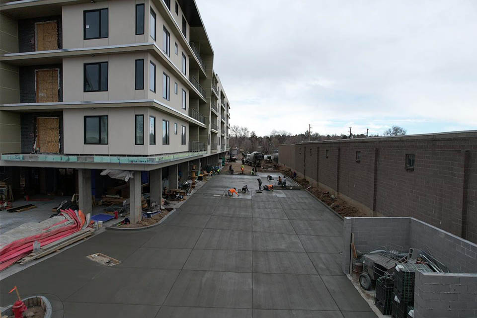 Construction workers are smoothing freshly poured concrete in a wide driveway beside a multi-story building under construction, with materials and equipment scattered around the site.
