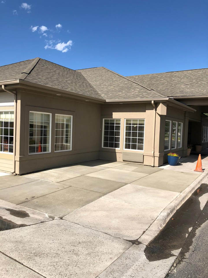 A beige building with multiple large windows, a sloped roof, and a paved sidewalk in front. There is a blue flower pot and orange traffic cones near the entrance. The sky is clear and blue.