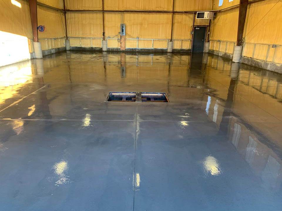 A large, empty warehouse with shiny, newly finished concrete floors. There is an open rectangular pit in the center of the floor, and the walls are lined with light wood paneling and structural beams.