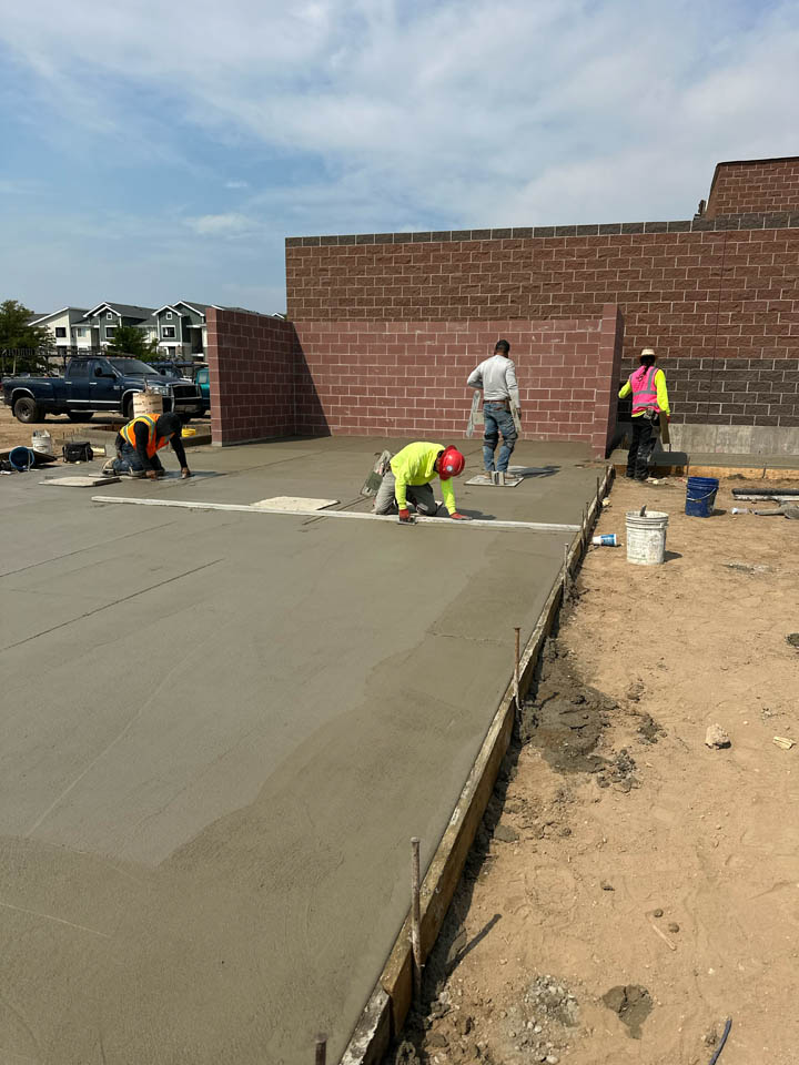 Four construction workers smooth and finish freshly poured concrete on a building foundation at a construction site, with brick walls and nearby buildings visible in the background.