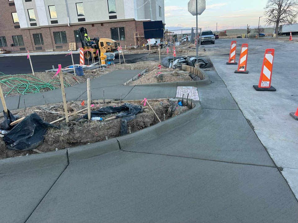 Freshly poured concrete sidewalks and curbs curve around a construction site with soil, stakes, and construction equipment visible. Orange safety barriers and a stop sign are in the background near a street and a building.