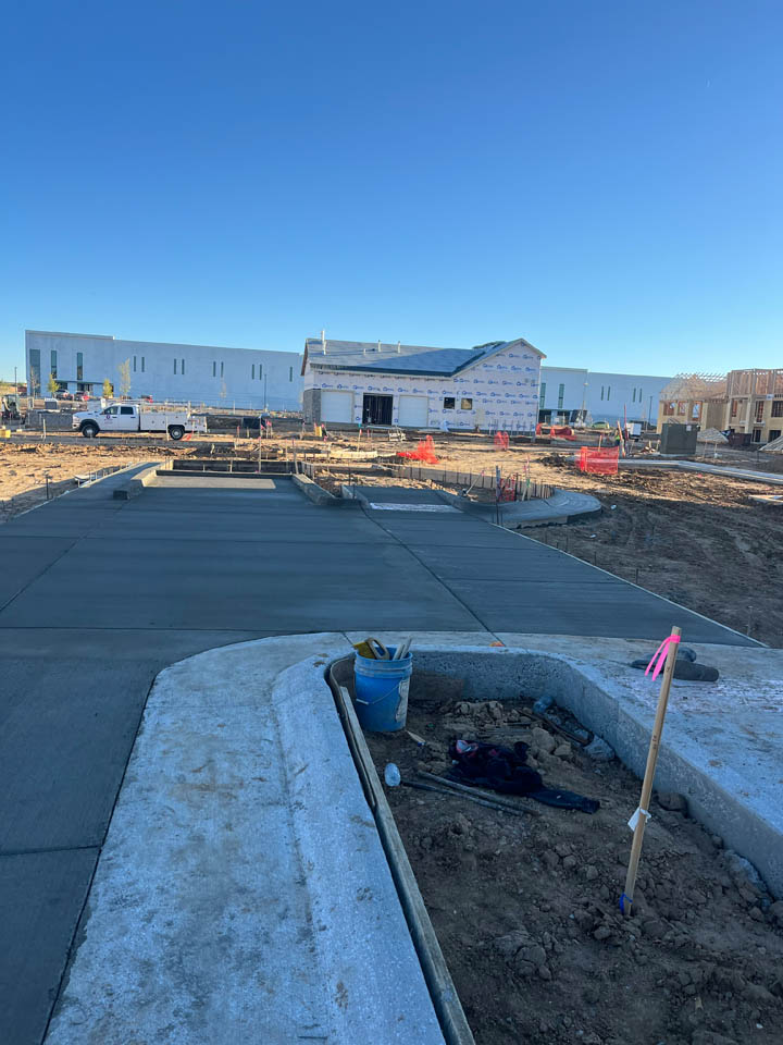 A construction site with freshly poured concrete, a blue bucket and tools in the foreground, and a partially built house with a white exterior in the background under a clear blue sky.
