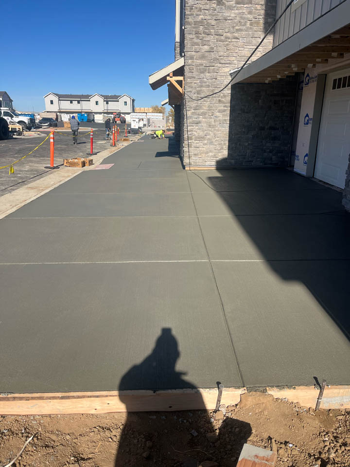 Freshly poured concrete driveway in front of a house under construction, with visible expansion joints. Construction workers and equipment are in the distance, and homes are visible in the background under a clear blue sky.