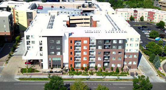 completed-commercial-project-2 Aerial view of a modern, multi-story apartment building with balconies, surrounded by trees, sidewalks, parked cars, and other buildings under a clear sky.