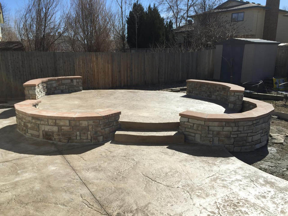 A backyard patio with a circular, stamped concrete surface and two curved stone benches with stone backs and flat tops. There are two steps leading up to the patio. A wooden fence and shed are in the background.