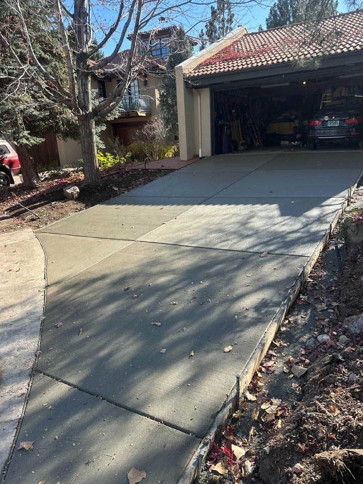 Freshly poured concrete driveway leads to an open garage with a car inside. Shadows of tree branches fall across the driveway. There are trees and landscaping around the house.