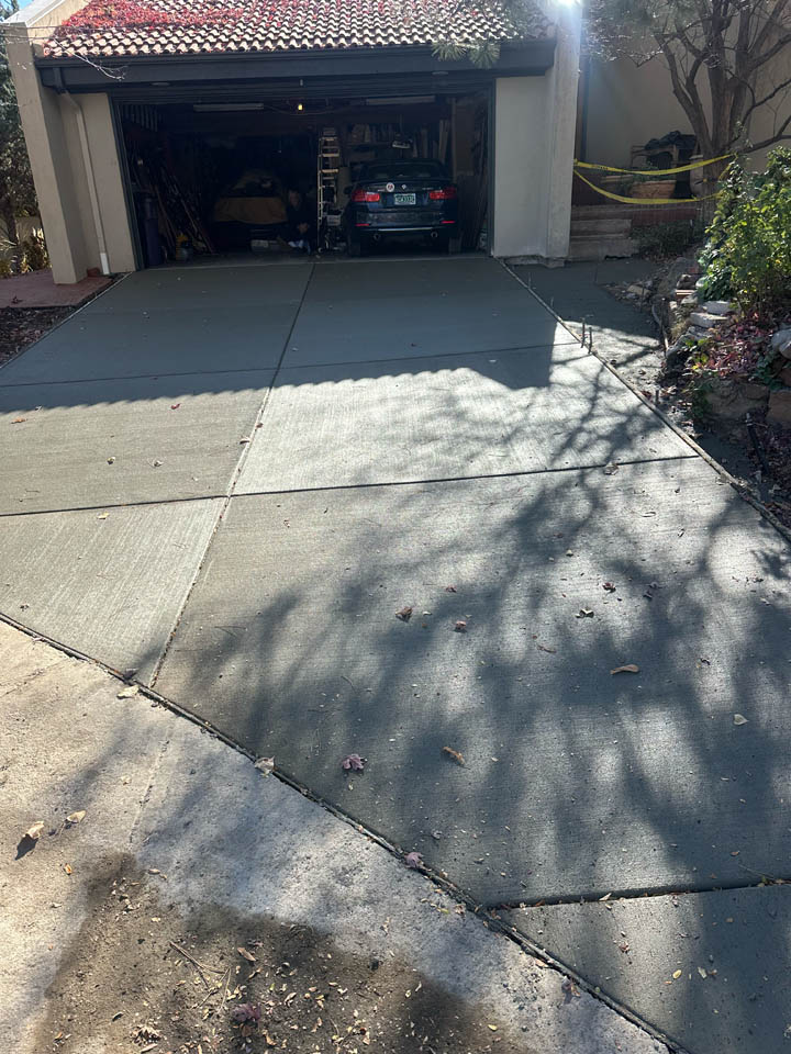 A driveway with freshly poured, smooth concrete leads to an open garage with a car parked inside. Sunlight casts tree shadows on the driveway, and yellow caution tape is visible on the right side.