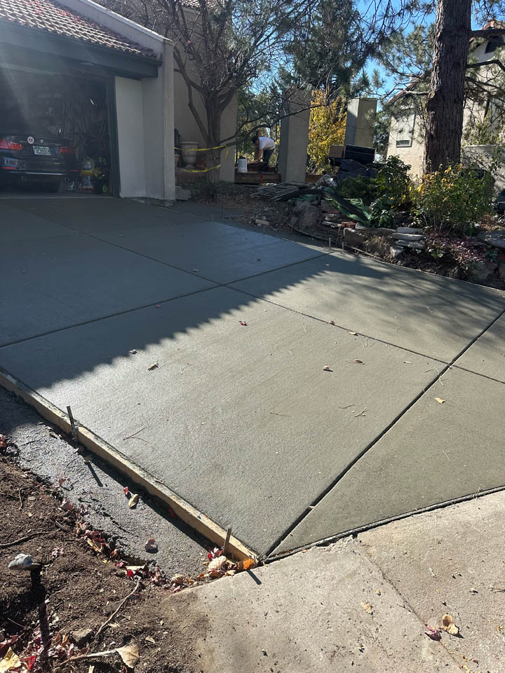 Freshly poured concrete driveway with visible seams, next to a garage with an open door. Yard debris and tools are nearby, and a person is working in the background near trees and landscaping.