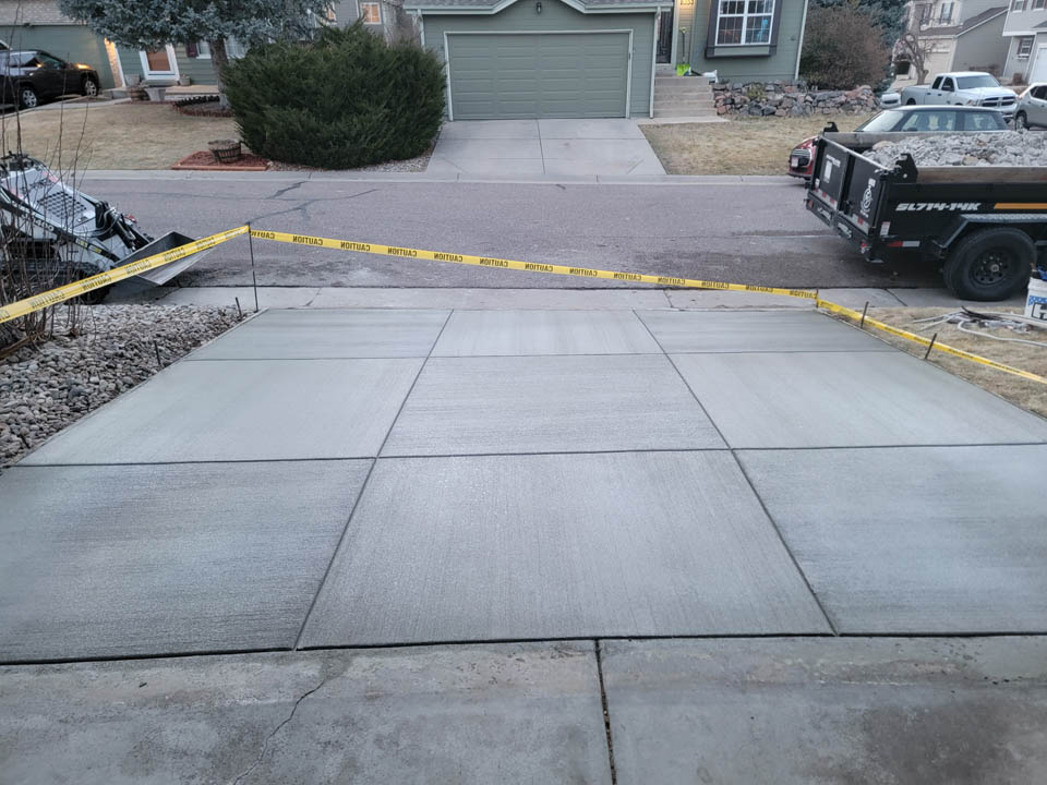 A newly poured concrete driveway sectioned into squares, roped off with yellow caution tape, in front of a house with a green garage door. A black utility trailer is parked nearby.