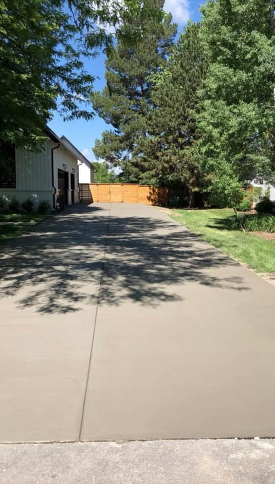 A newly paved concrete driveway leads to a building with white siding and a tan wooden fence at the end, surrounded by green grass, trees, and landscaping on a sunny day.