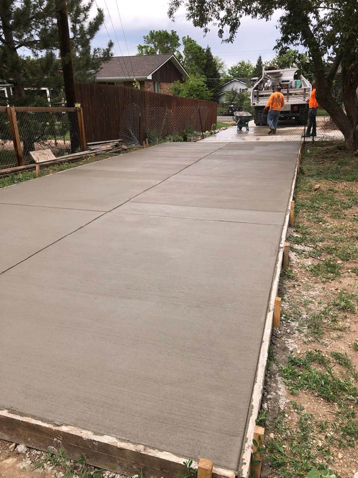 Freshly poured and smoothed concrete driveway with workers in safety vests in the background. The driveway is bordered by wooden forms, with houses, trees, and fencing visible alongside.
