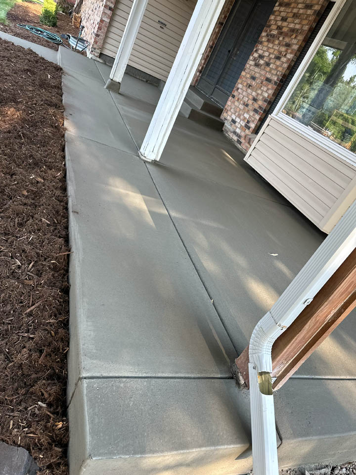 Freshly poured and smoothed concrete sidewalk and porch area in front of a house with beige siding and brick exterior, next to a new garden bed with mulch and a white downspout.