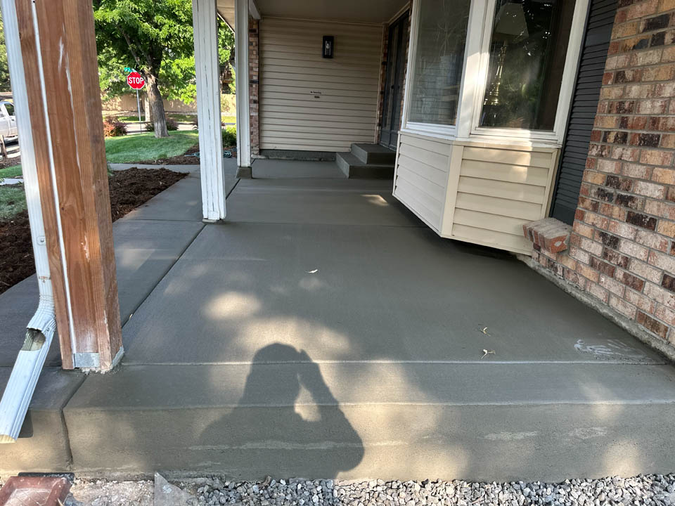 Freshly poured concrete covers a front porch and walkway of a house with brick and siding. A shadow of a person taking the photo is visible on the concrete, and there are a few small leaves on the surface.