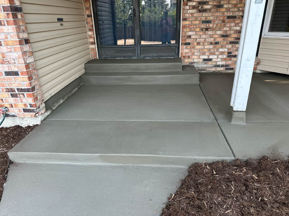 Freshly poured and smoothed concrete steps and walkway lead to a glass door entrance of a brick and siding building; mulch lines the edge of the walkway.