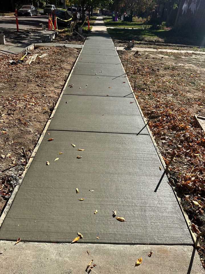 Freshly poured concrete sidewalk with a few scattered leaves on top, surrounded by dirt and some autumn leaves. Safety cones and caution tape are visible in the background near the street.