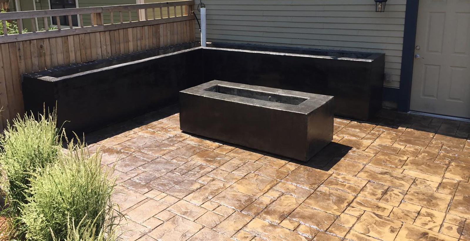 A patio with stamped concrete flooring, a rectangular concrete planter in the center, L-shaped raised concrete planters along the fence and wall, and green shrubs in the foreground.