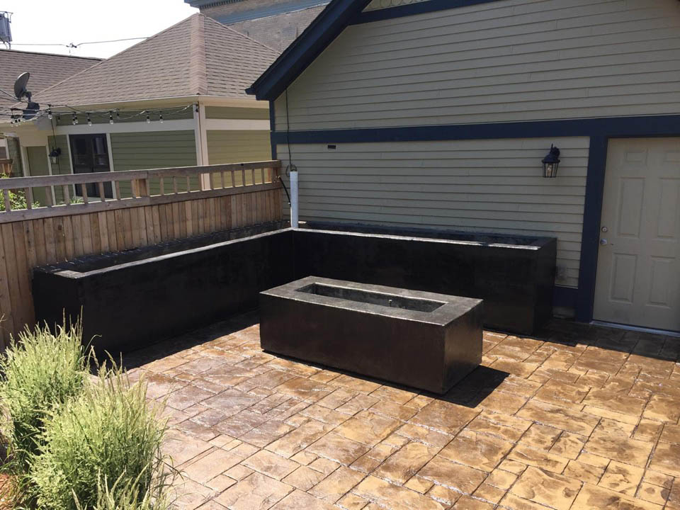 A paved patio with a wooden fence, featuring a rectangular raised planter in the center and an L-shaped black raised planter along the corner near a house with light siding and a white door.
