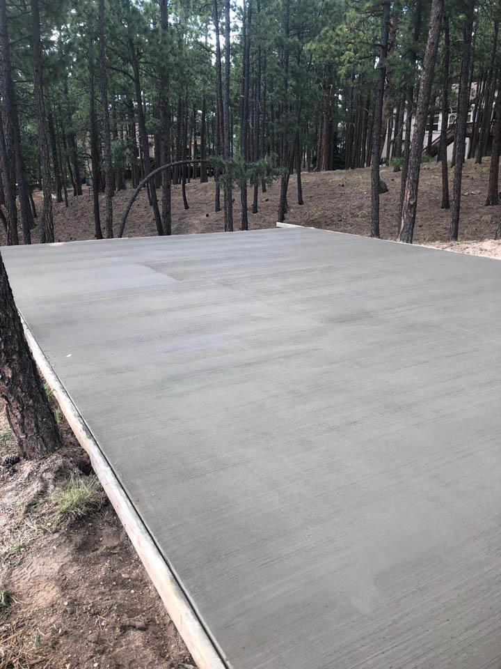 A freshly poured and smoothed concrete slab sits in a forested area surrounded by tall pine trees, with no structures yet built on the slab.