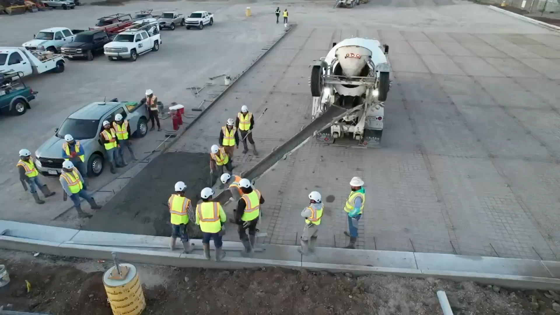 Construction workers in safety vests and helmets guide wet concrete being poured from a cement mixer truck onto a roadway, while other workers and vehicles are nearby on the construction site.